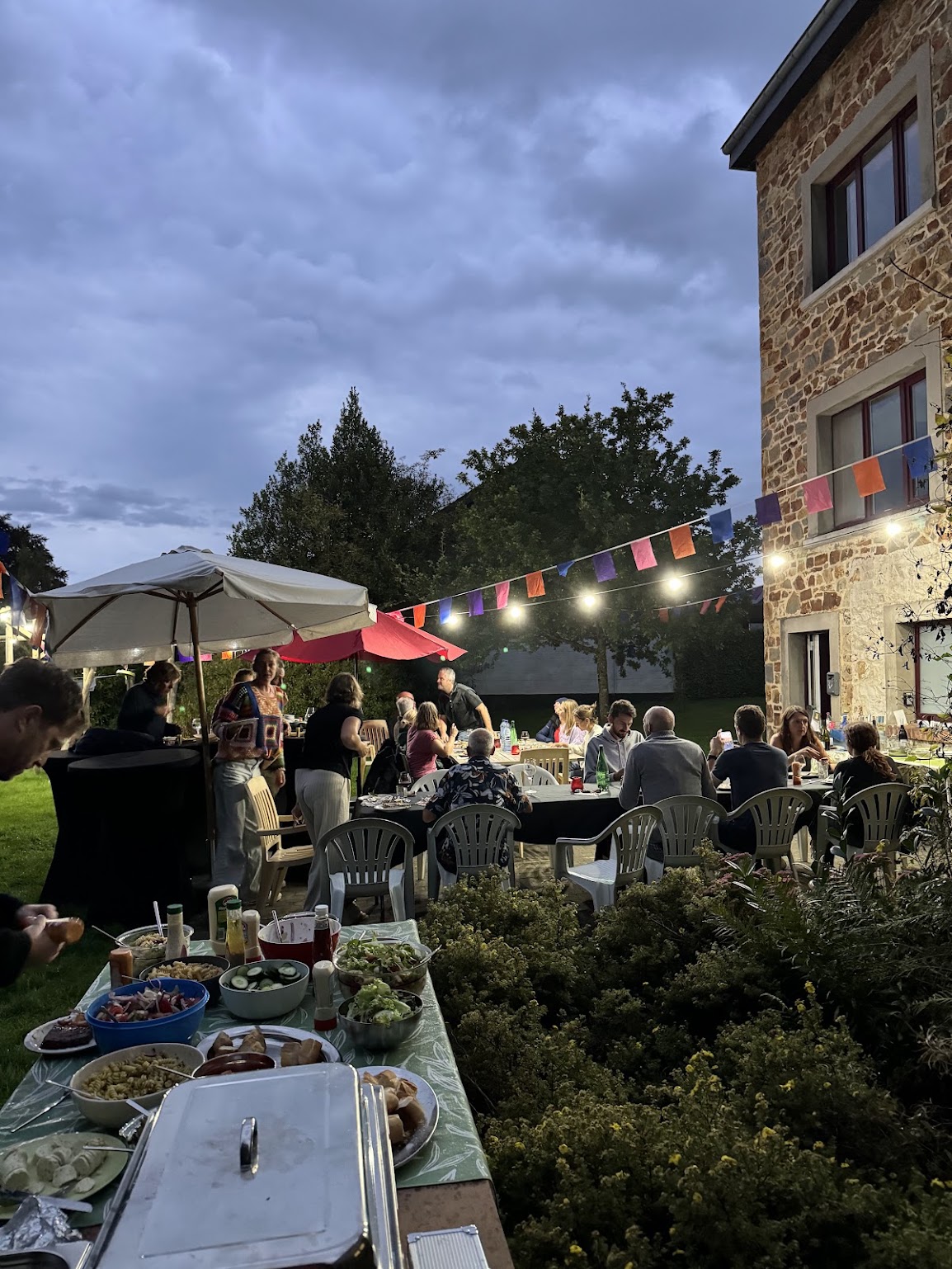 Vrienden aan een lange tafel in de tuin van Ferme au Bois bij avond